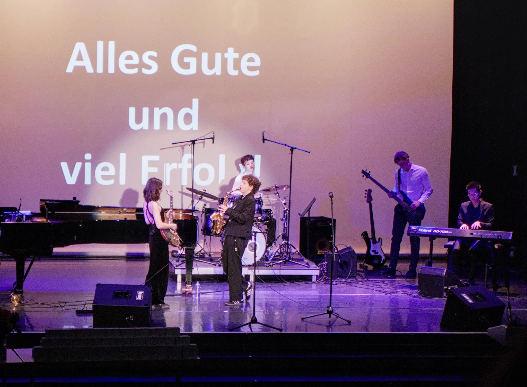 Die ganze Band auf einer Bühne mit Flügel, im Hintergrund eine Leinwand mit "Alles Gute und viel Erfolg"